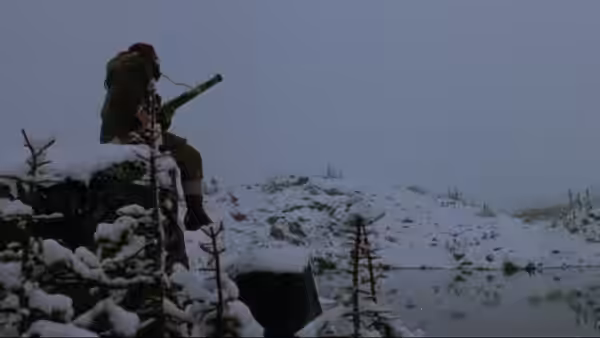 A bundled figure sits on a snow-covered ridge, holding a rifle