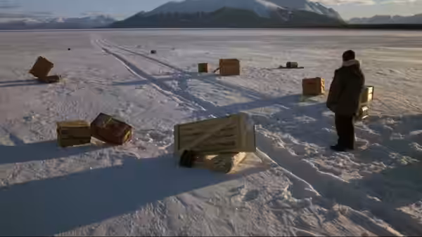 A bundled figure stands among crates scattered across a snowy plain