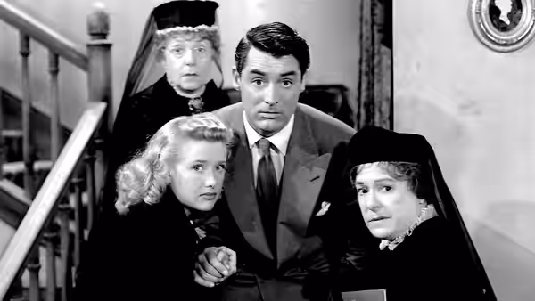 Mortimer Brewster (Cary Grant) stands between two veiled women on a staircase landing
