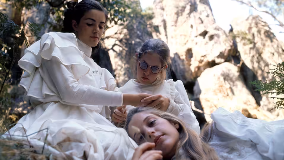 Three girls in white dresses gather closely among sunlit rocks outdoors