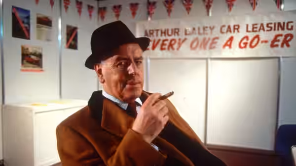 Arthur Daley (George Cole) sits in his car leasing office, holding a cigarette