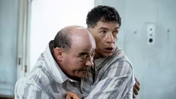A younger man in a striped shirt holds an older man close as he looks anxiously aside