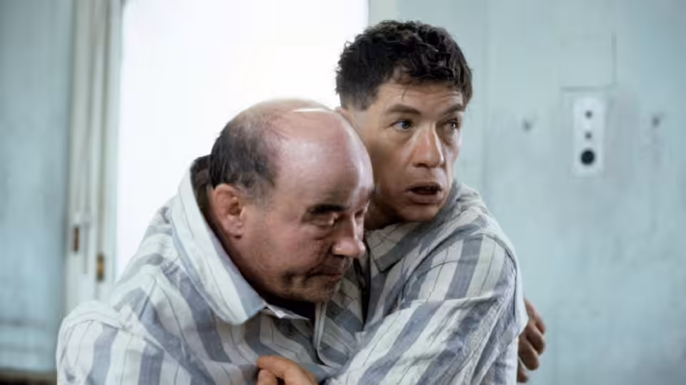 A younger man in a striped shirt holds an older man close as he looks anxiously aside