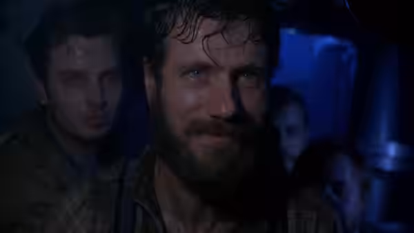 A bearded sailor watches tensely as crew members gather behind him
