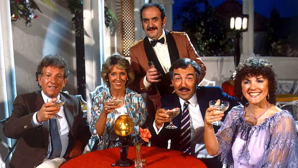 Four guests raise drinks around a table as a waiter stands behind them