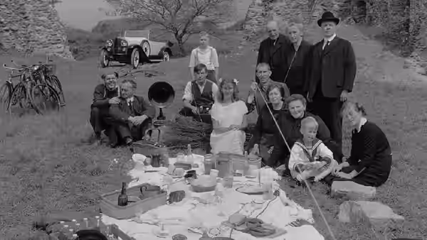 A family gathers around a picnic spread beside stone ruins and a parked car