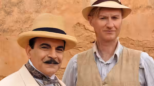 Hercule Poirot (David Suchet) stands beside a man in a vest against a sandstone wall