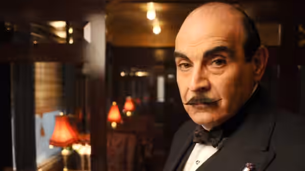 Hercule Poirot (David Suchet) stands in a dimly lit wood-paneled room