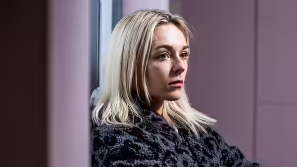 A blonde woman in a dark coat stands beside a doorway