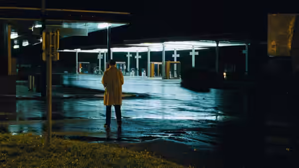 A man in a long coat stands alone at a brightly lit gas station at night