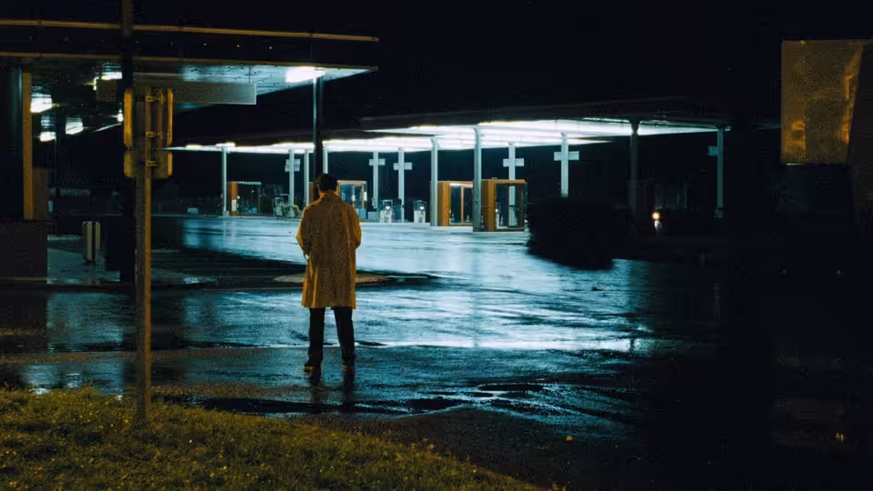 A man in a long coat stands alone at a brightly lit gas station at night