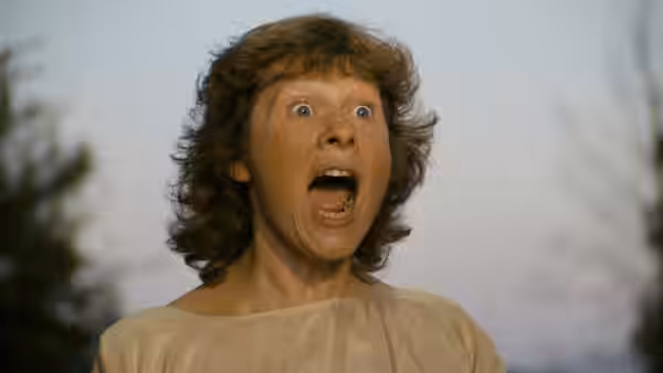 A woman in a pale blouse stares ahead with her mouth open