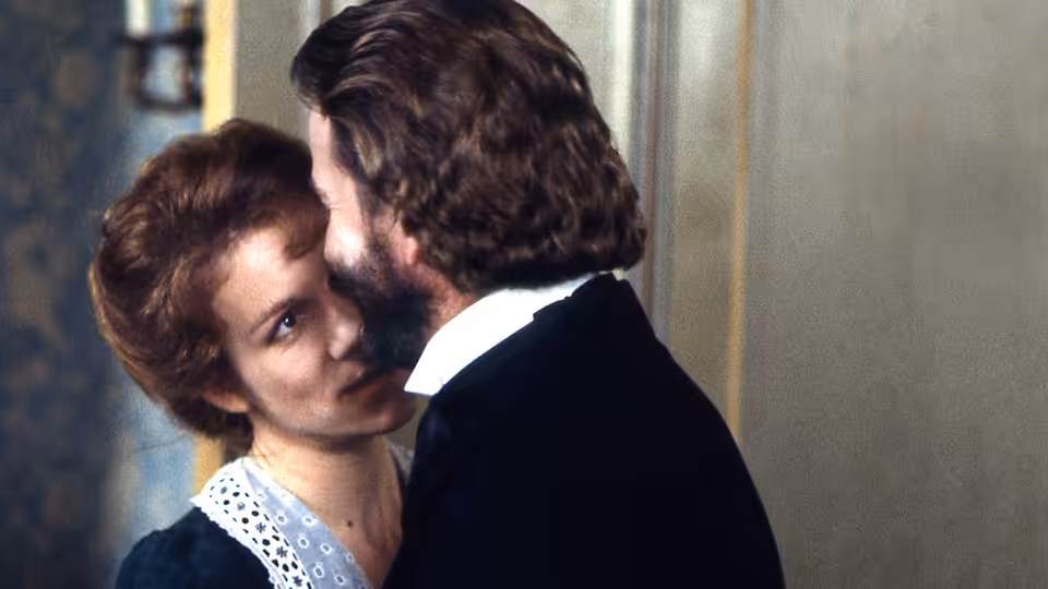 Nora Helmer (Juliet Stevenson) gazes up at Torvald Helmer (Trevor Eve) as he leans close beside her