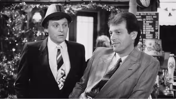 Two suited men stand in a pub, one wearing a trilby and tie