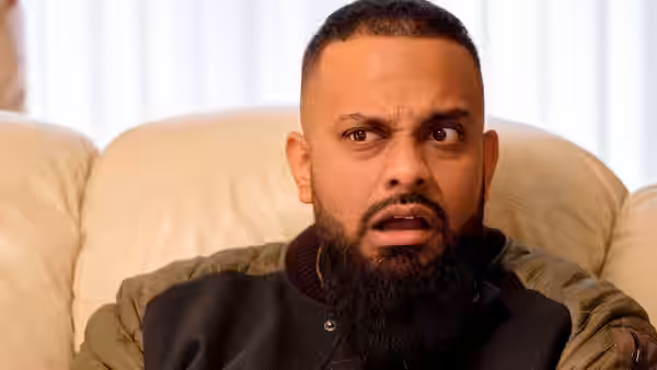 Mobeen (Guz Khan) reacts with a startled expression while seated on a sofa