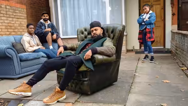 Mobeen (Guz Khan) reclines in an armchair outside a house as three men watch