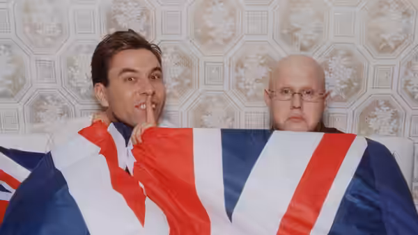 Two men sit beneath a Union Jack blanket, one picking his nose