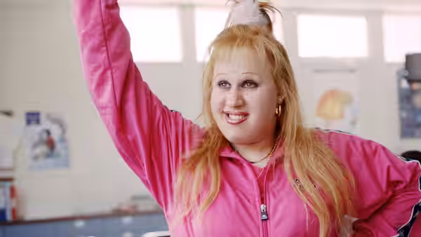 Fat Pat (Joann Condon) raises an arm in a bright pink tracksuit