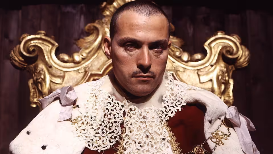 A man in a lace-trimmed coat sits before an ornate gilded throne
