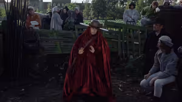 A red-cloaked figure gestures before a group gathered by a wooden fence