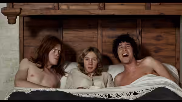 A woman lies between two bare-chested men in bed