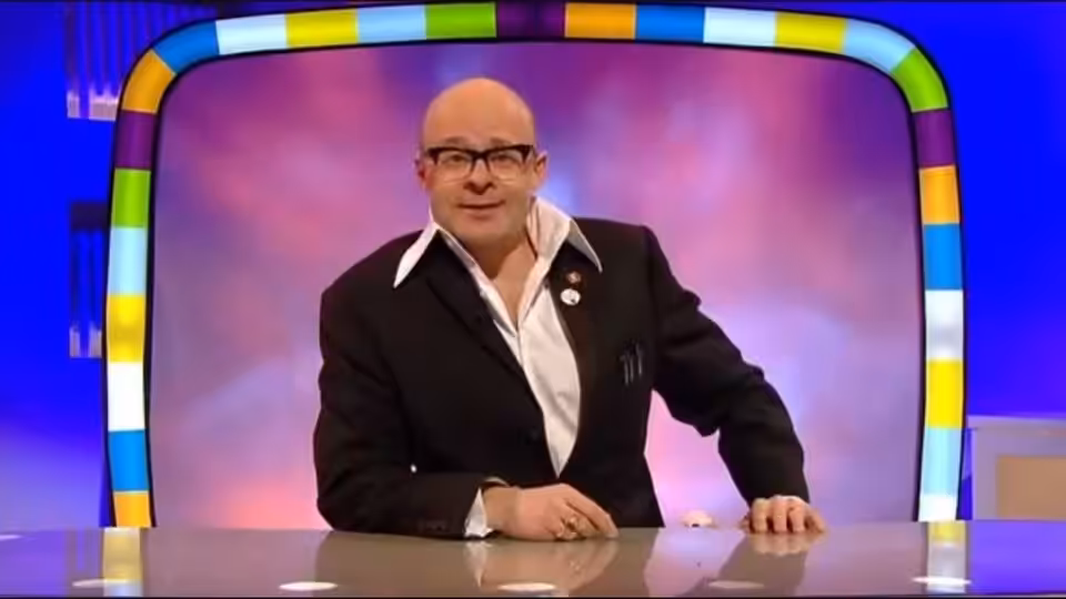 Presenter (Harry Hill) leaning on a desk beneath a multicoloured arch