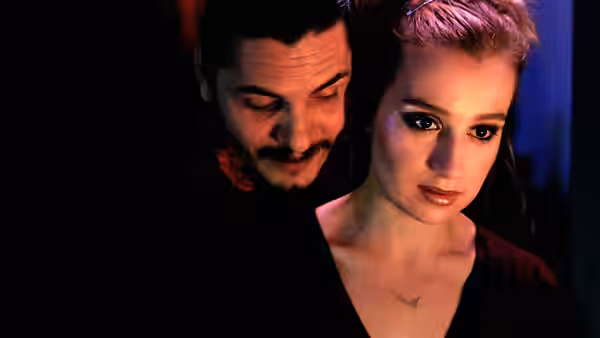 A man leans close behind a young woman in a dimly lit room