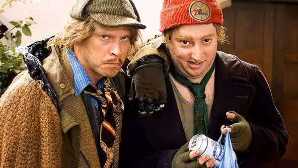 Two men in winter clothes stand together, one holding a can of beer
