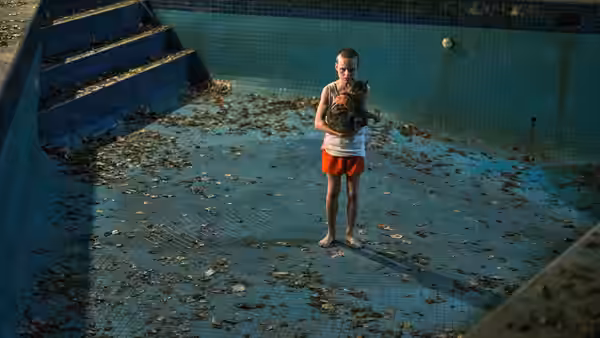 A boy stands barefoot in a leaf-strewn pool cradling a cat