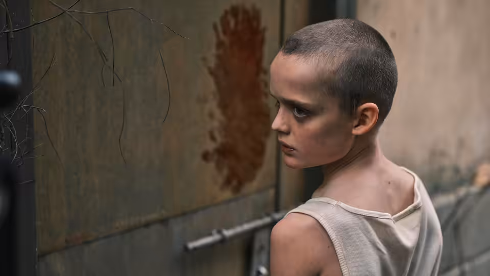 A boy with a shaved head stands against a rusted metal wall