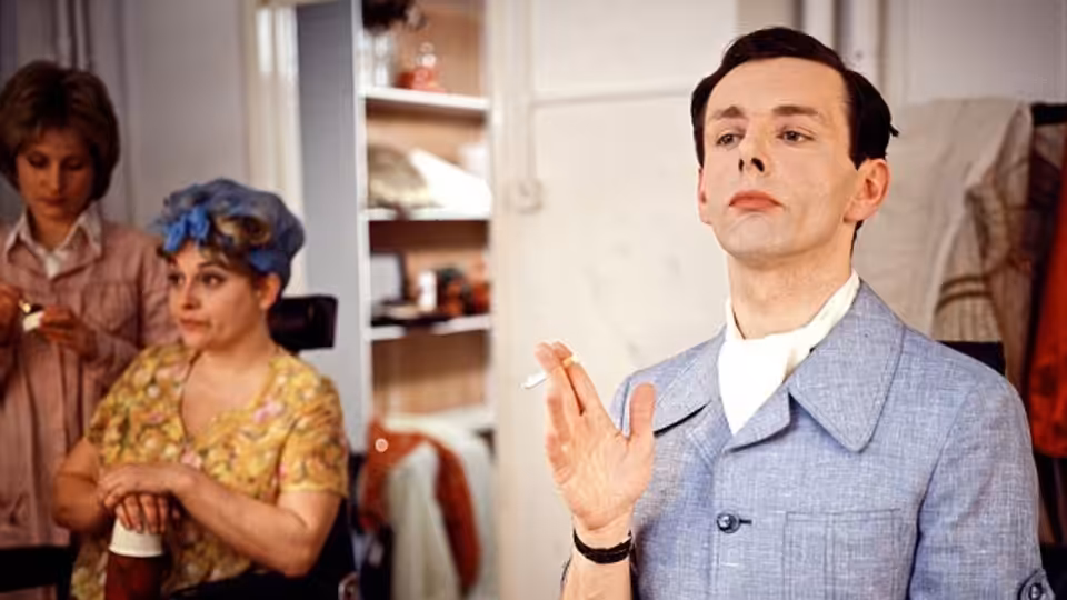 Kenneth Williams (Michael Sheen) gestures in a blue suit as two women stand behind him