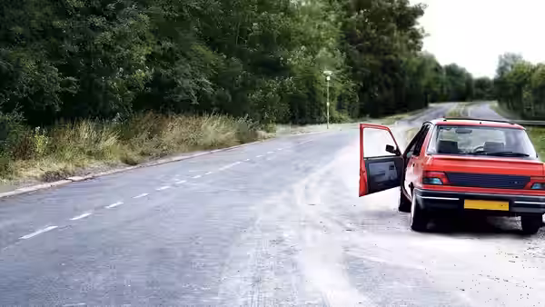 A red hatchback sits on a deserted country road with its driver's door open