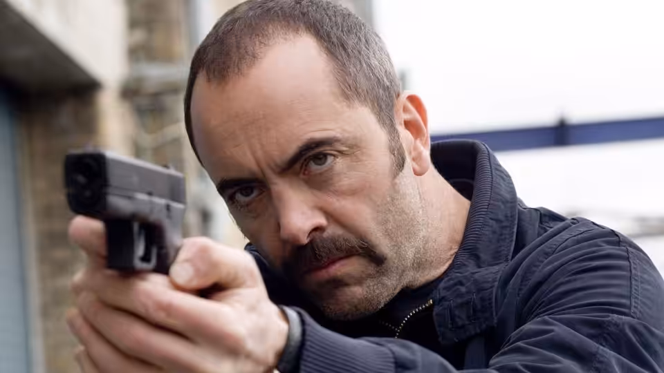 DS Tommy Murphy (James Nesbitt) aiming a handgun outdoors beside a brick building