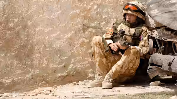 A soldier sits against a stone wall, holding his rifle in dusty combat gear
