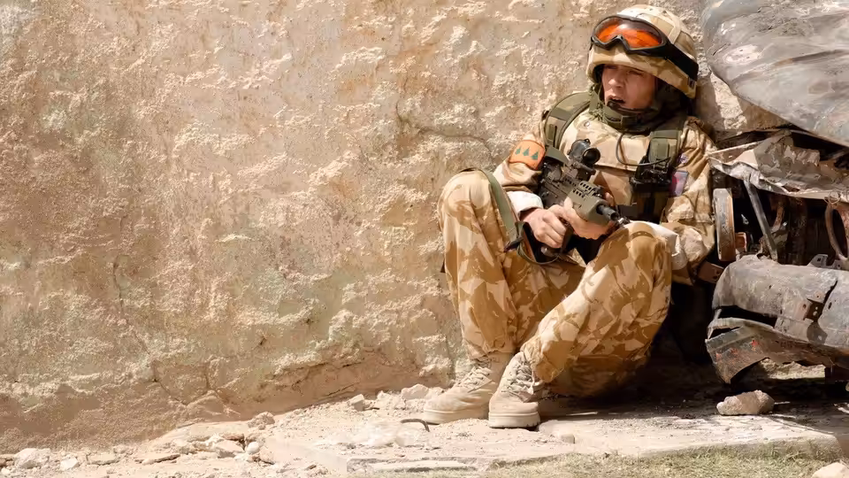 A soldier sits against a stone wall, holding his rifle in dusty combat gear