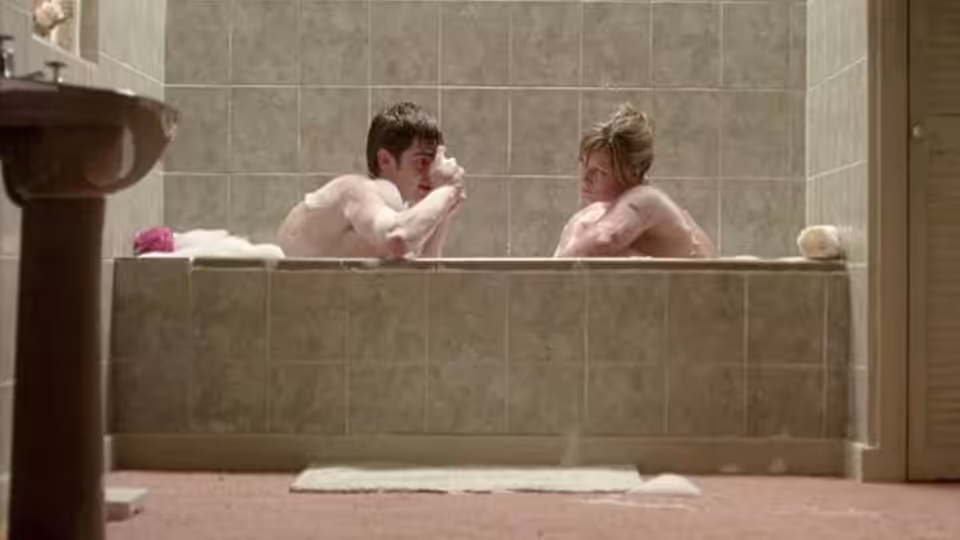 A young man and woman sit facing each other in a bathtub