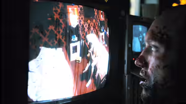 A bloodied man watches a television showing a figure inside the house