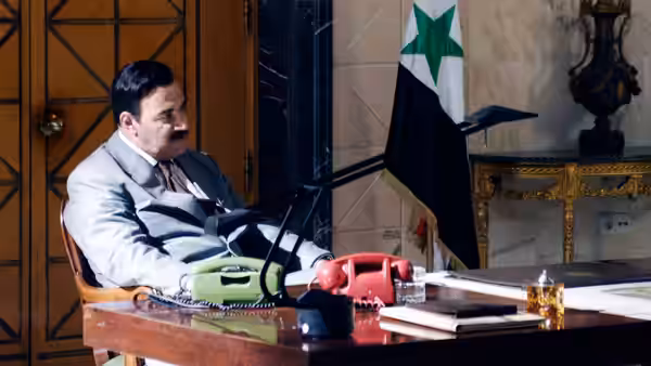 Saddam Hussein (Yigal Naor) seated at his desk beside the Iraqi flag