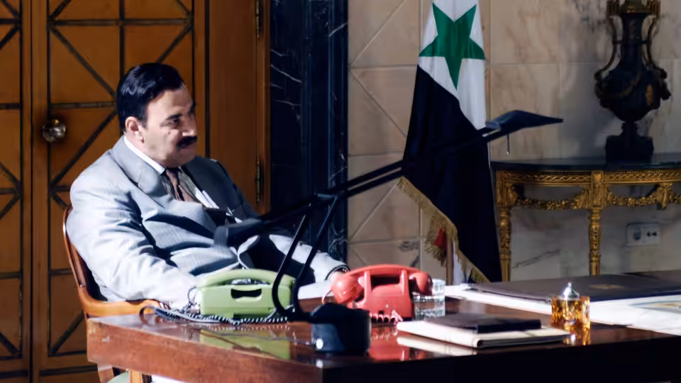 Saddam Hussein (Yigal Naor) seated at his desk beside the Iraqi flag