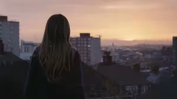A girl stands with her back to the camera, looking over city rooftops at sunset