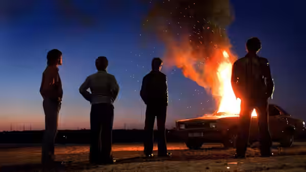 Four young men watch a car burn beside a rural road at dusk