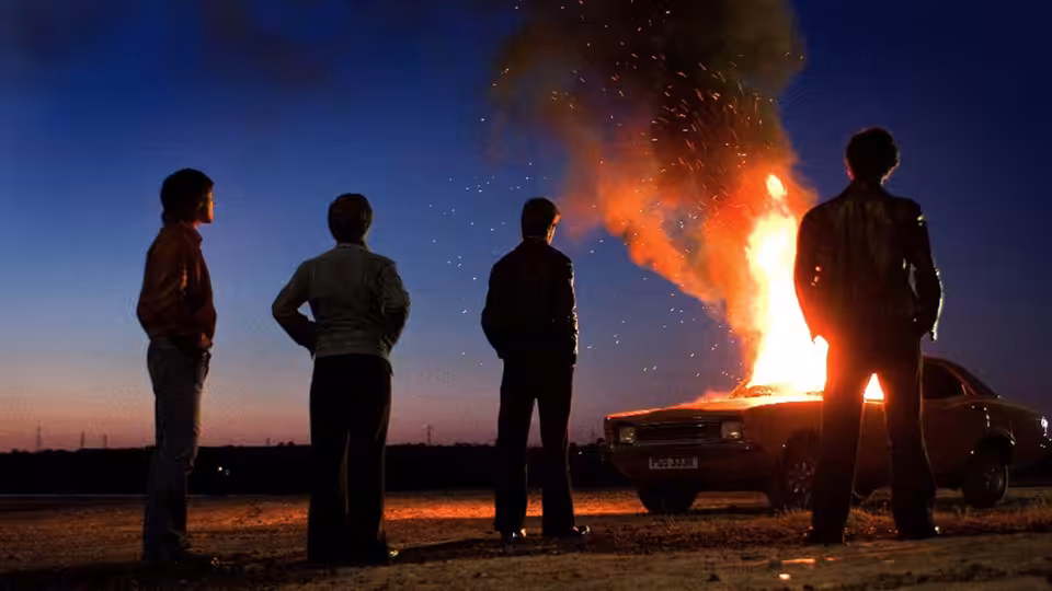 Four young men watch a car burn beside a rural road at dusk