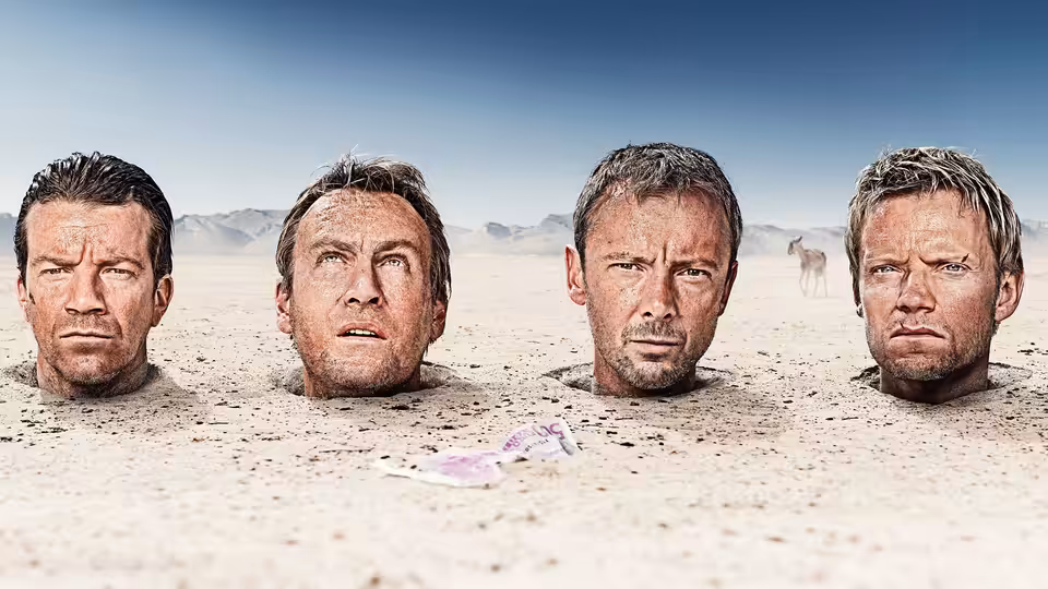 Four men buried up to their necks in desert sand, staring ahead