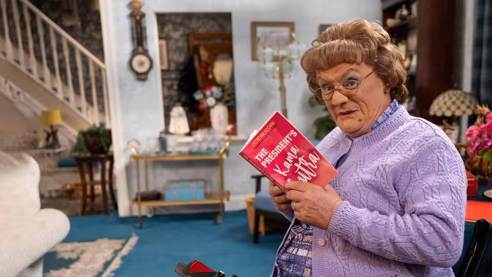 Agnes Brown (Brendan O'Carroll) holds a red book in her living room
