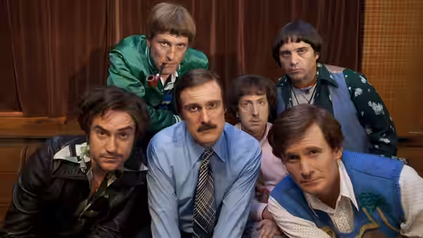 Six men in varied suits and costumes lean together in a wood-paneled room, facing forward