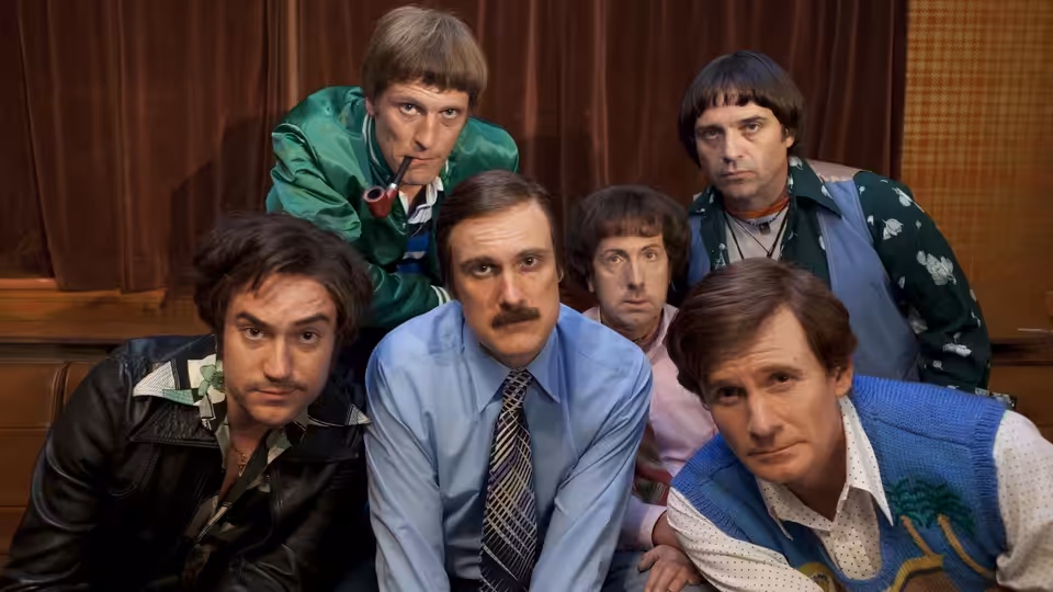 Six men in varied suits and costumes lean together in a wood-paneled room, facing forward