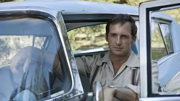 A man sits in an open car doorway in a wooded area
