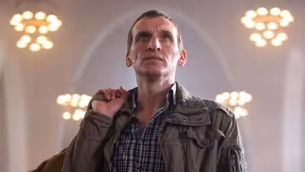 A man in a checked shirt and jacket looks upward beneath bright ceiling lights