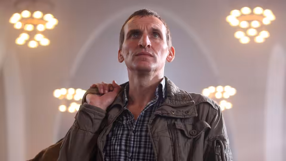 A man in a checked shirt and jacket looks upward beneath bright ceiling lights