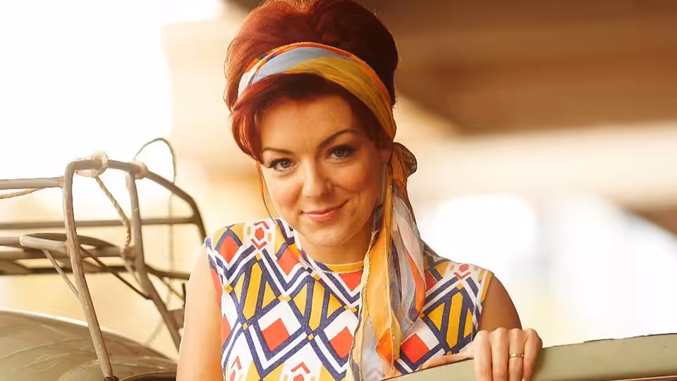 Charmian Biggs (Sheridan Smith) in a colorful patterned dress and headscarf, leaning beside a car